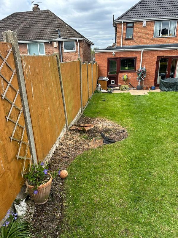 Lawn alongside a tall wooden fence with a small cut stump and bare soil where the tree had been