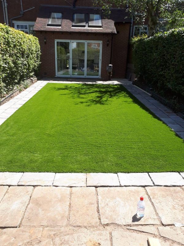 Artificial lawn bordered by paving slabs in a back garden facing a brick house with bi-fold doors