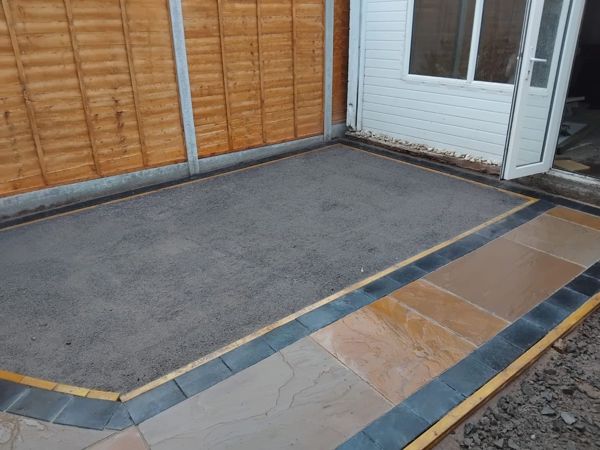 Part-laid patio of orange sandstone-look slabs with dark block edging and a grey stone base beside new fence panels