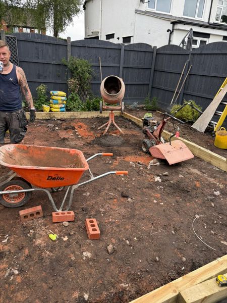 Cleared back garden with an orange wheelbarrow, cement mixer and rotavator on bare earth beside a dark fence