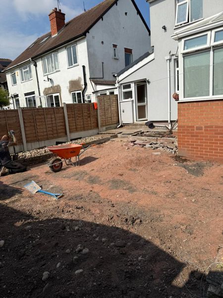 Graded reddish ground with an orange wheelbarrow and a pebble strip along a timber fence