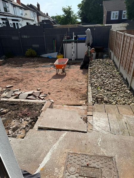Sand spread across a garden with old paving slabs, a drain cover and a worker near the pebble border