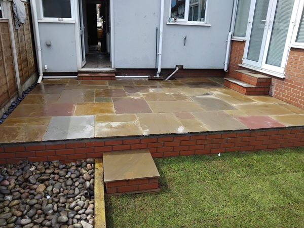 Wide view of a raised patio with brick front and small step beside a pebble border and new lawn