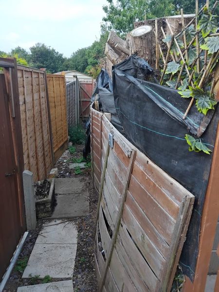 Narrow side passage with a leaning weathered fence, black netting and cut logs stacked above it