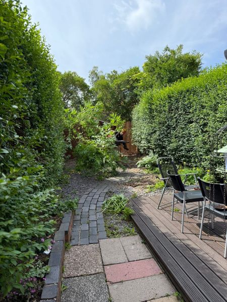 Brick-edged garden path between tall hedges, with cut green foliage and a worker at the far end near a decked seating area