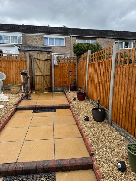 New close-board fence with a gate, paved path edged in brick, and gravel borders with pots and a statue