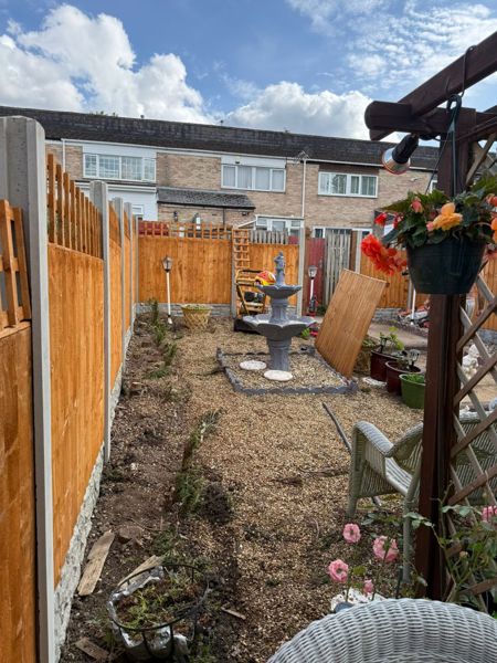 New fence panels on concrete posts beside a bare soil strip, with a fountain and a leaning panel in the gravel garden