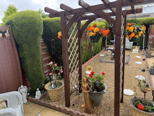 Wooden pergola with orange hanging baskets, pots and statues, and a clipped conifer hedge behind