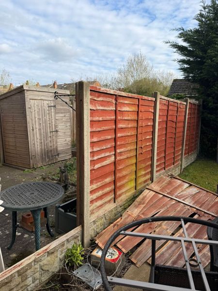 Faded red overlap fence with one panel collapsed onto a patio near a garden table and shed